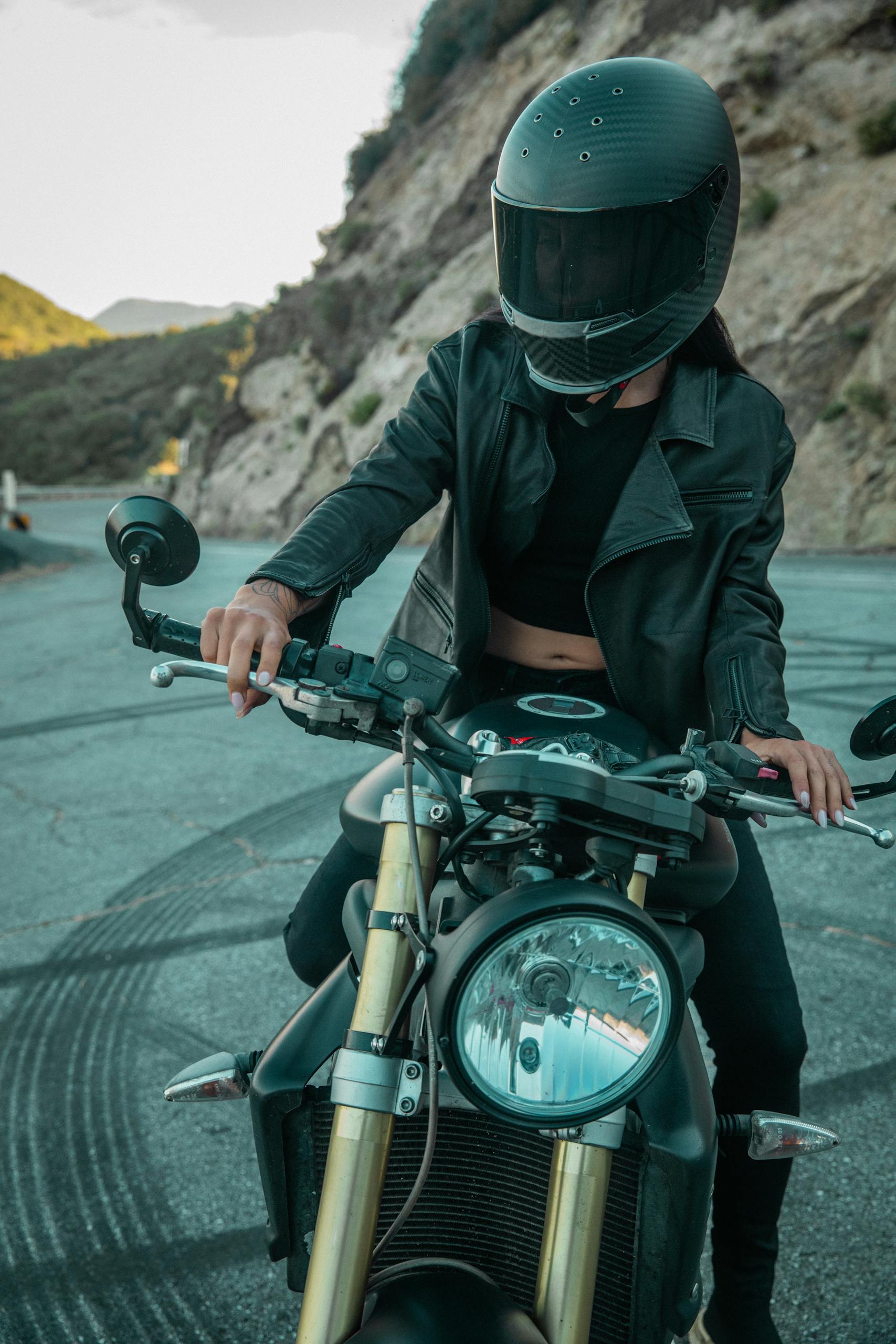 A biker in black leather gear poses confidently on a motorcycle on a winding mountain road.